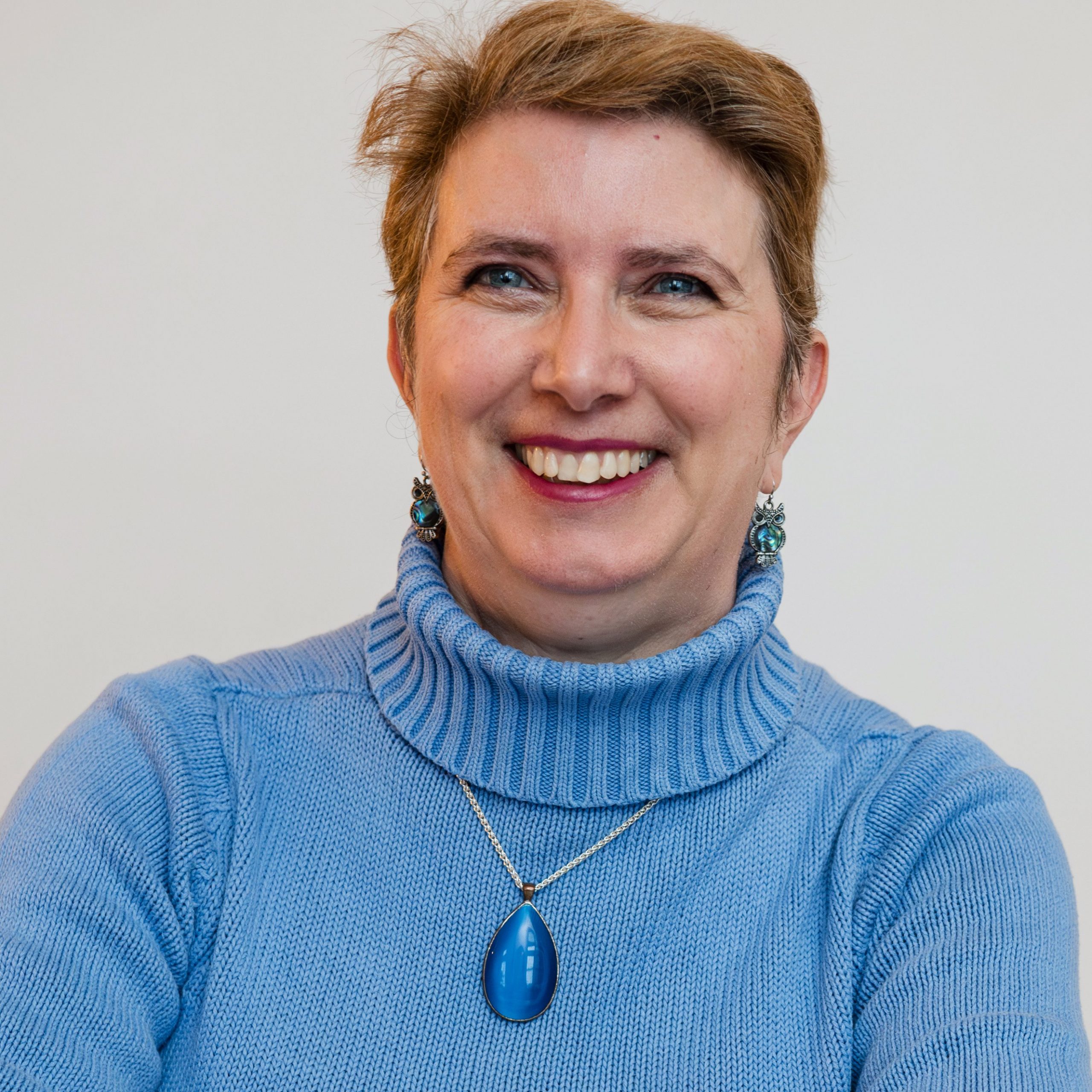 Headshot of Della Hudson FCA smiling and wearing a blue jumper and pendant necklace. Della has short, brown hair in a pixie cut and likes to think she looks younger than her 50 something years